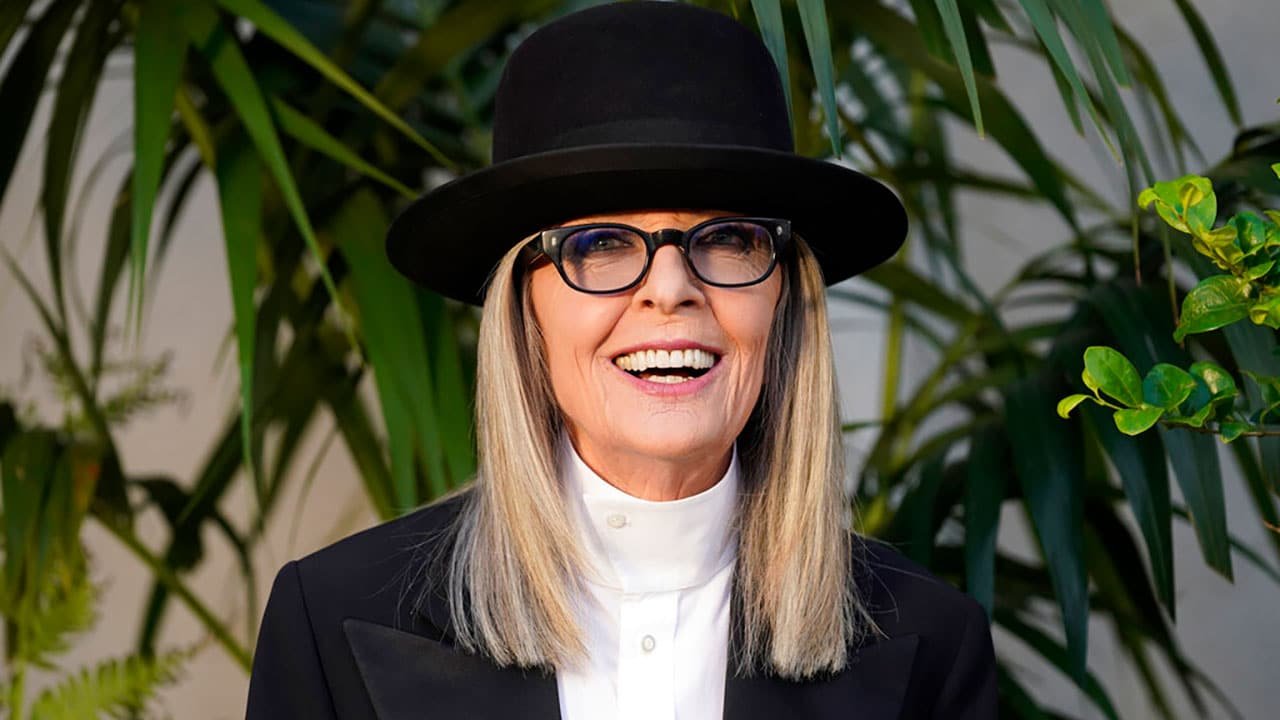 Diane Keaton wearing a black hat and glasses, smiling, with long straight hair, and dressed in a black suit with a white shirt, standing outdoors with green plants in the background.