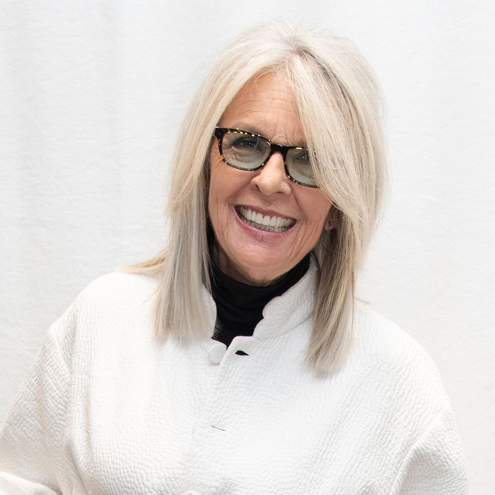 Diane Keaton with long gray hair wearing glasses and a white textured jacket smiles against a plain white background.