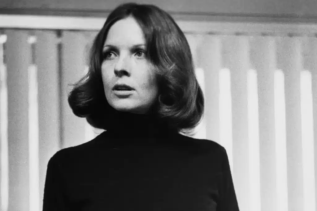 Black and white photo of Diane Keaton with shoulder-length hair looking off-camera, wearing a dark turtleneck sweater against a background with vertical blinds. Here, she played Kay in the Godfather movies.
