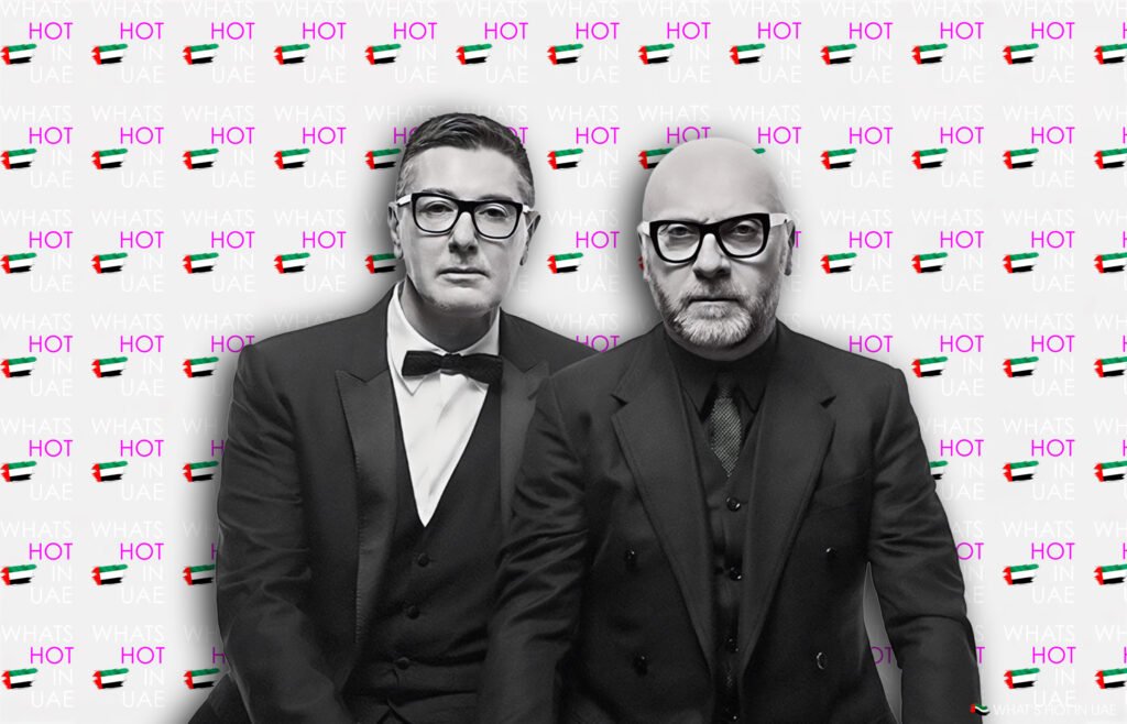 The Unvarnished History of Dolce & Gabbana: Genius, Controversy, and Legacy Two men in black suits and glasses stand in front of a backdrop with repeated text "WHAT'S HOT IN UAE" and images of the UAE flag.