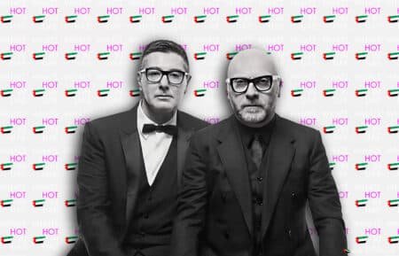 Two men in black suits and glasses stand in front of a backdrop with repeated text "WHAT'S HOT IN UAE" and images of the UAE flag.