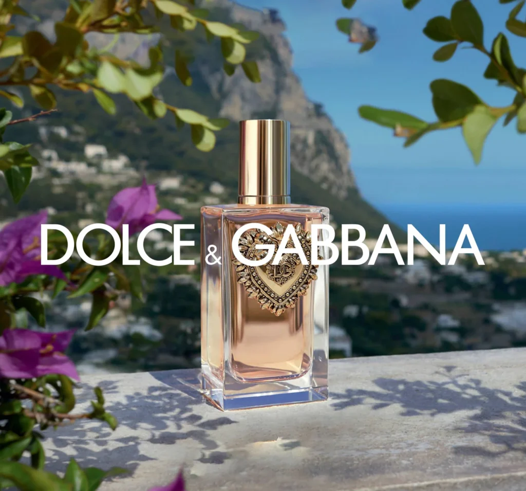 A Dolce & Gabbana perfume bottle with a gold cap and ornate heart design on a ledge, surrounded by vibrant purple flowers and overlooking a scenic coastal landscape.
