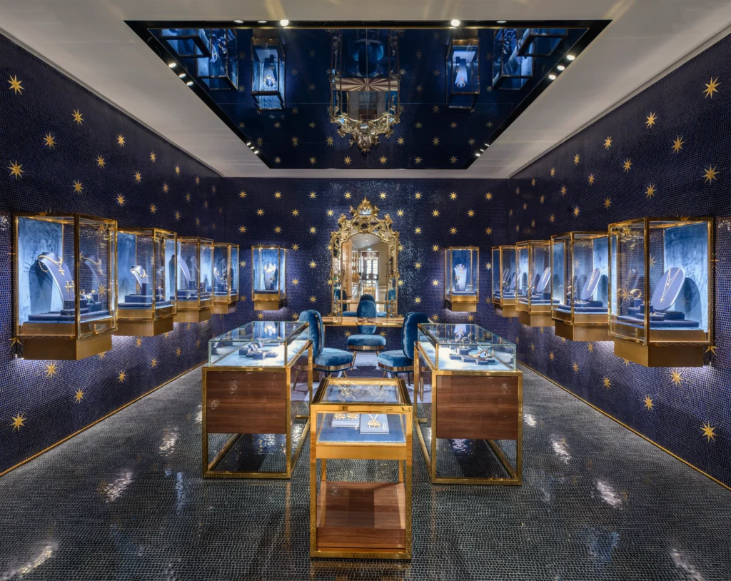 The History of Dolce & Gabbana: A luxurious jewelry store with dark blue walls adorned with golden star patterns, featuring glass display cases showcasing necklaces and other jewelry. There is a large ornate mirror on the far wall and plush blue velvet chairs. The floor is glossy and reflective, adding to the opulent atmosphere.