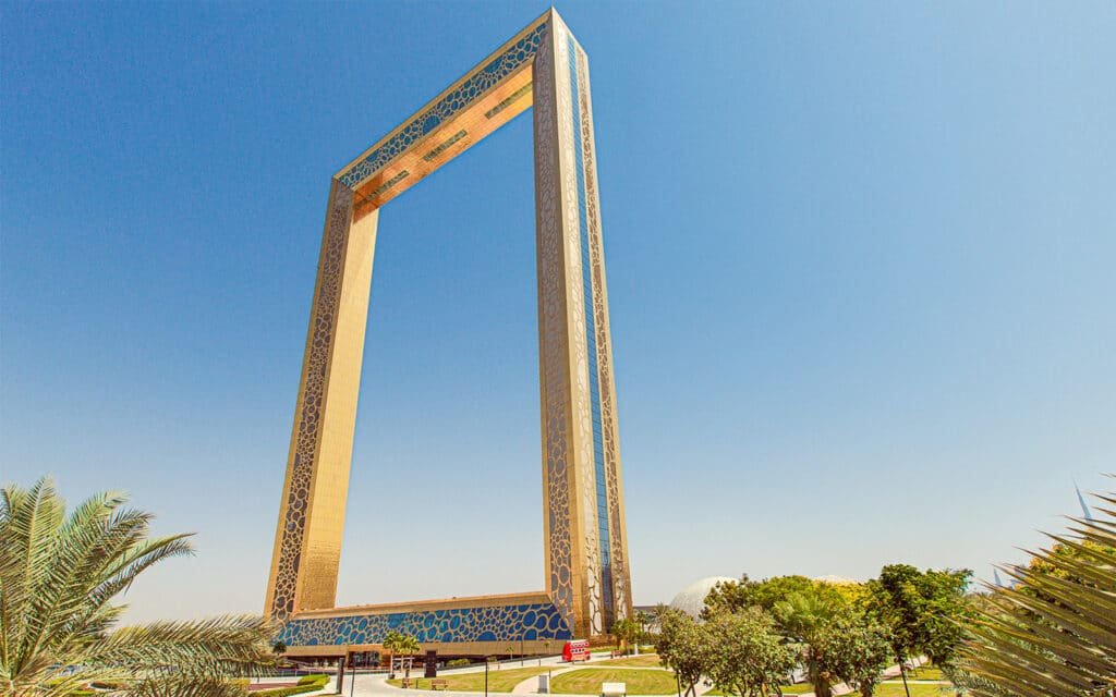 Tourist Spots in Dubai:  A tall, rectangular frame-shaped structure with ornate patterns, set against a clear blue sky, surrounded by a landscaped park with palm trees and greenery. This is the Dubai Frame.