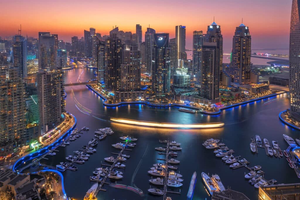 Aerial view of a city marina at dusk, with tall skyscrapers and illuminated waterways surrounded by multiple docked yachts, under a vibrant orange and purple sky.