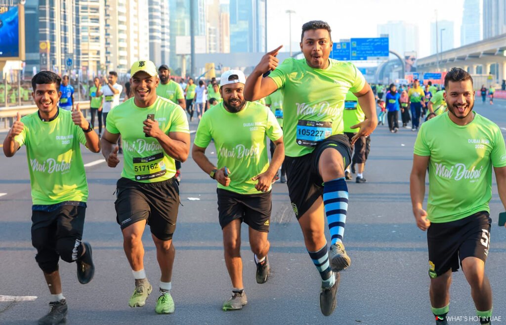 Dubai Run 2025: A group of five men in bright green t-shirts participate in a running event, joyfully moving and posing on a city street with tall buildings in the background. One man is leaping in the air with a playful expression, while others give thumbs up and smile at the camera. All are wearing running shorts and athletic shoes, and some have race bibs with numbers.