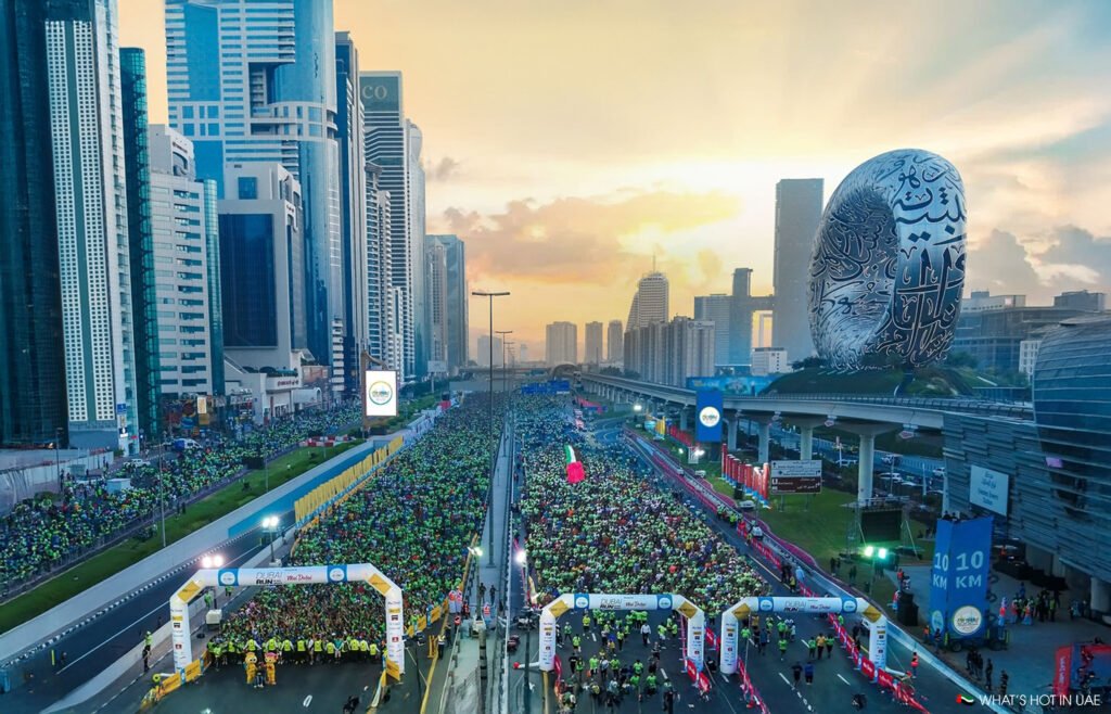 A large crowd of people participates in a marathon on a wide boulevard surrounded by tall skyscrapers, with a distinctive, egg-shaped building featuring Arabic script in the background. The sun is setting, creating a colorful sky.