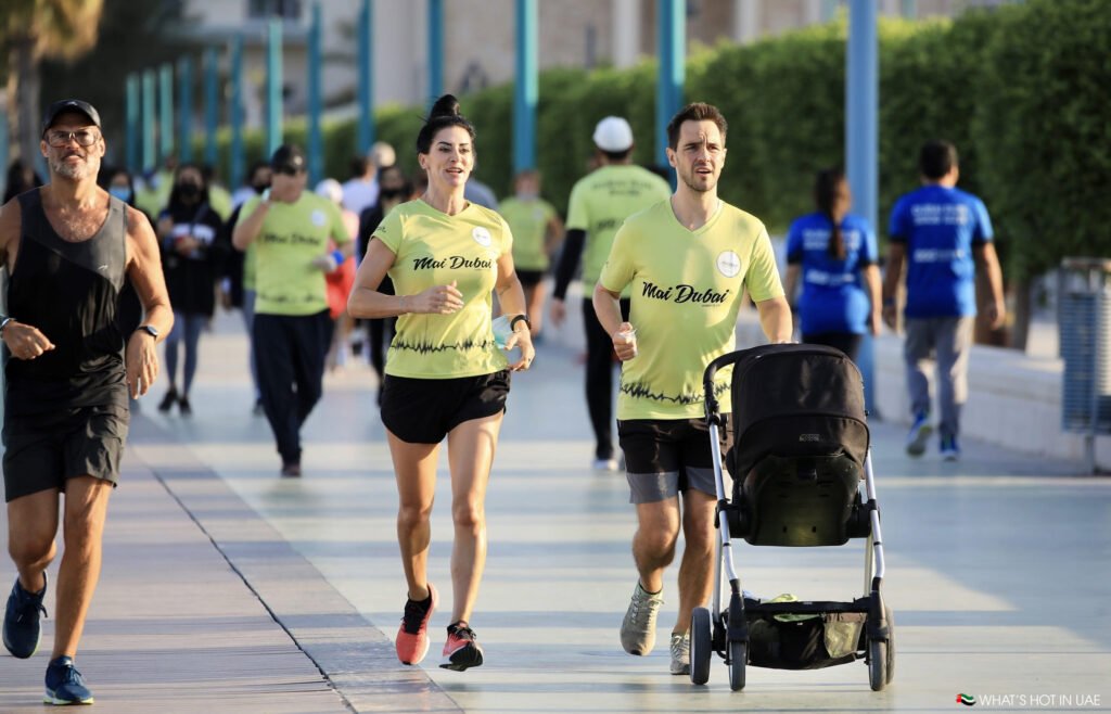 Dubai Run 2025: A group of people jogging on a paved path, with two individuals wearing yellow shirts labeled 'Mai Dubai' and a man pushing a baby stroller.