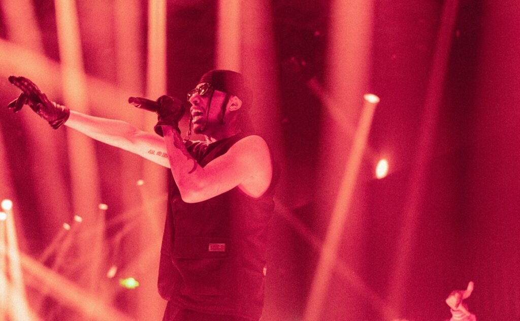 EZOW on stage, wearing sunglasses and a headscarf, singing into a microphone with dynamic red lighting and outstretched arm.