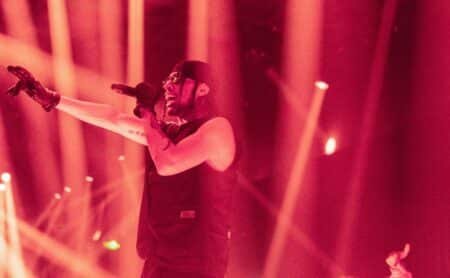 EZOW on stage, wearing sunglasses and a headscarf, singing into a microphone with dynamic red lighting and outstretched arm.