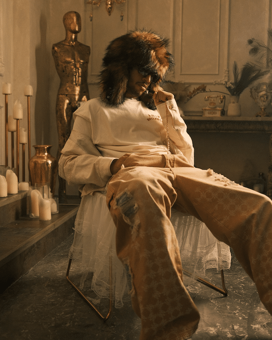 EZOW seated in a stylish room wearing a fur hat, sunglasses, and casual clothing, surrounded by decorative candles and a gold statue.