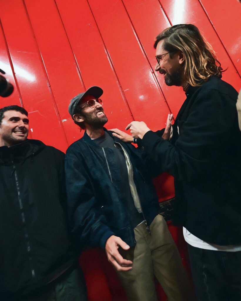 Three men standing together, smiling and laughing in front of a red wall.