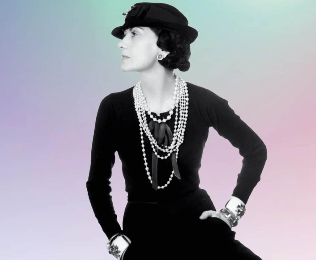 A woman in a black outfit and hat, adorned with several strands of pearl necklaces, stands against a softly colored gradient background.