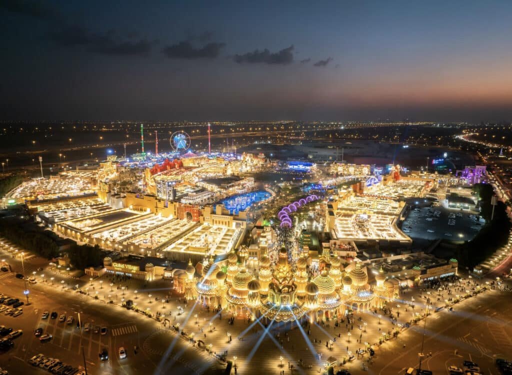 Aerial view of Global Village in Dubai at night, showcasing brightly lit buildings and attractions, including a Ferris wheel and various themed structures, amidst a bustling crowd and parking areas.