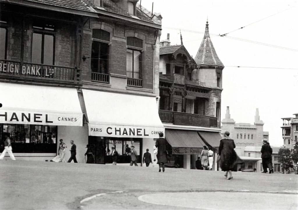A vintage black and white photograph showing a street scene with a Chanel store in Cannes. Several people are walking by, and the architecture is characterized by classic European-style buildings with pointed roofs and decorative facades. The store has awnings displaying the Chanel name prominently.