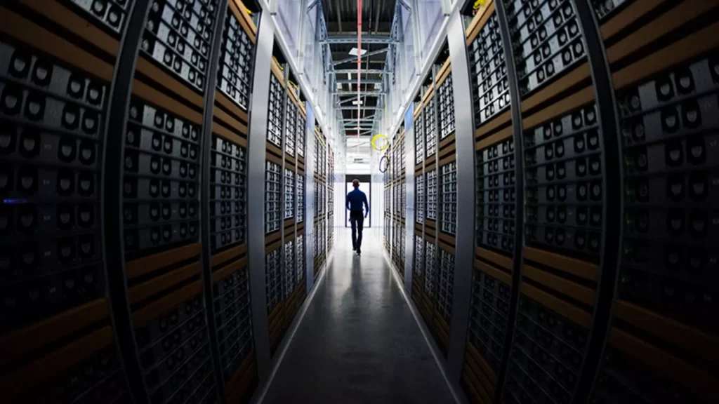 A person walking down an aisle in a data center, surrounded by rows of server racks with blinking lights.