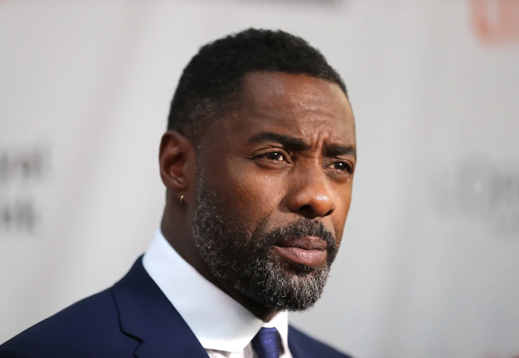 Idris Elba wearing a navy blue suit and white shirt, with a sharp focus on his serious expression and well-groomed beard, against a blurred background.