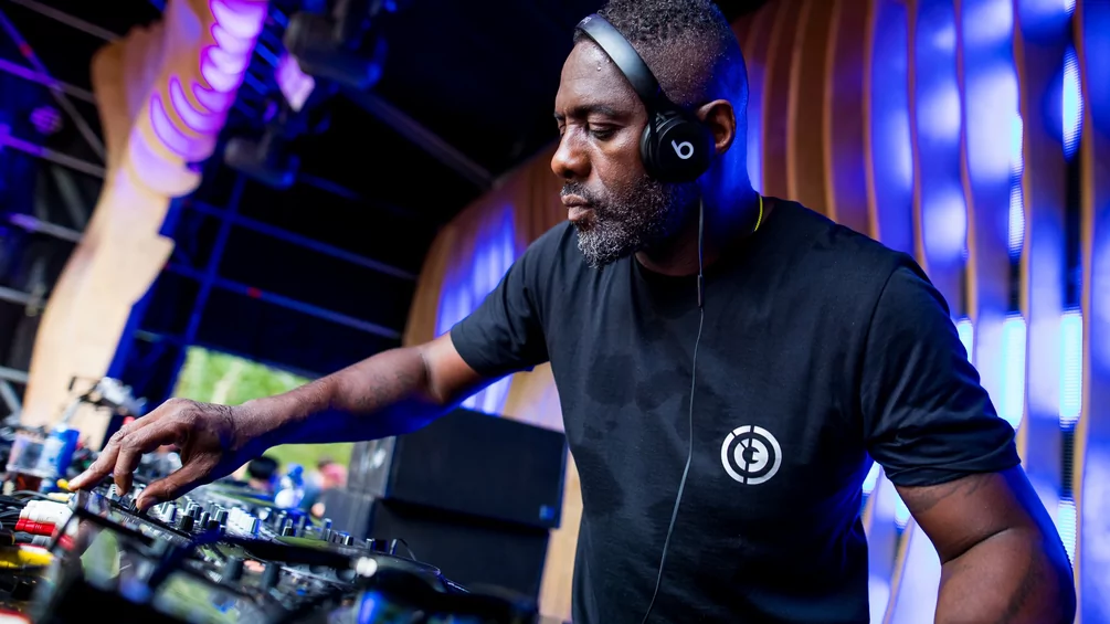 Idris Elba wearing headphones is focused on mixing music on a soundboard at an indoor event with colorful lighting in the background.