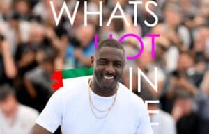 Idris Elba in a white t-shirt smiles at an event, with the words "WHAT'S HOT IN UAE" overlaid in large white and pink letters, and a brushstroke of the UAE flag colors in the background.