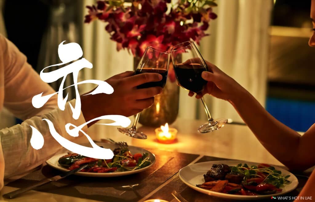 A romantic dinner setting with two people clinking wine glasses over candlelight, surrounded by plates of food and flowers, with stylized white text overlay.