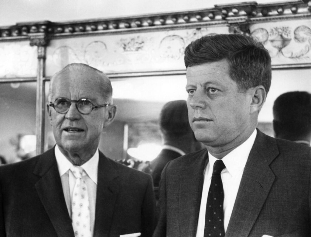 Two men in suits stand in front of a decorative mirror, one wearing glasses and the other with a neat hairstyle, both with serious expressions.