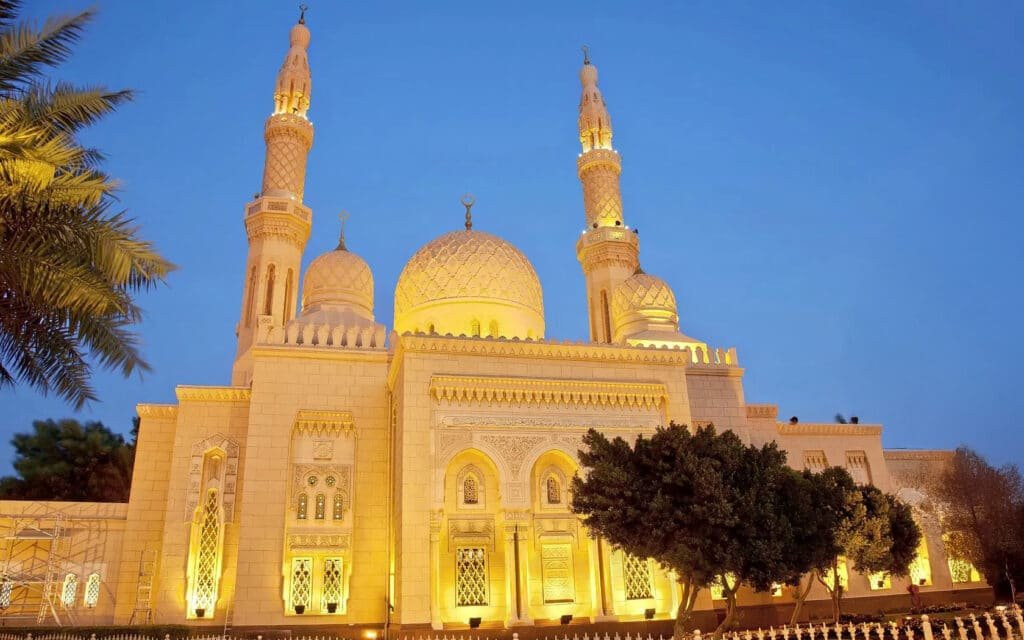 Tourist Spots in Dubai: Jumeirah mosque with illuminated golden architecture and three minarets and domes, set against a clear evening sky with palm trees nearby.
