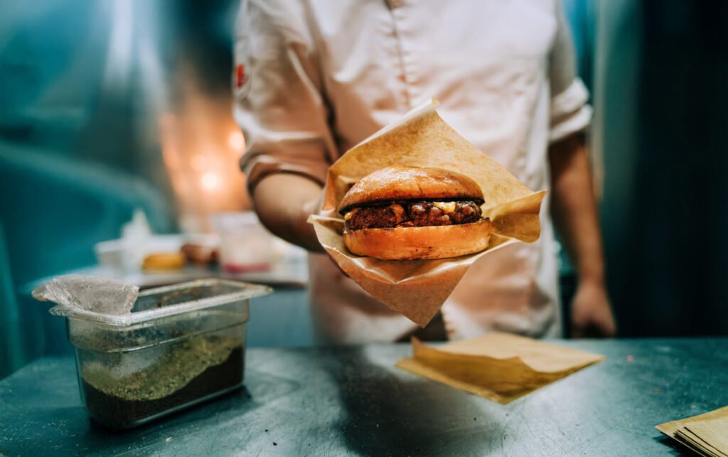 A chef holds a freshly prepared burger wrapped in brown paper, with a plastic container of seasoning on a metal counter in an industrial kitchen setting.