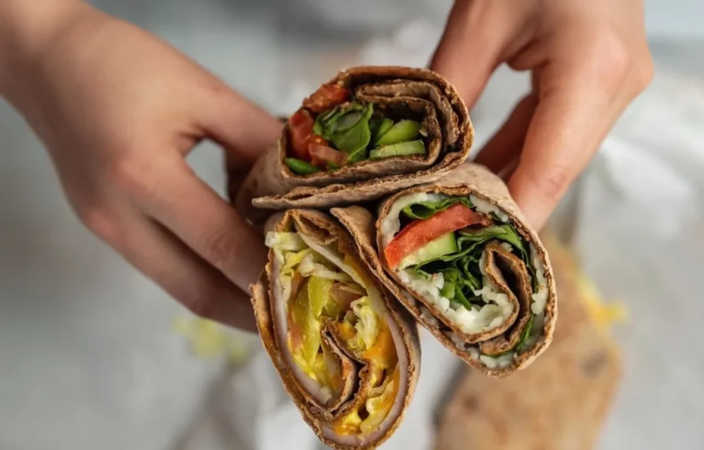 Close-up of two hands holding three whole wheat wraps filled with mixed vegetables and greens.