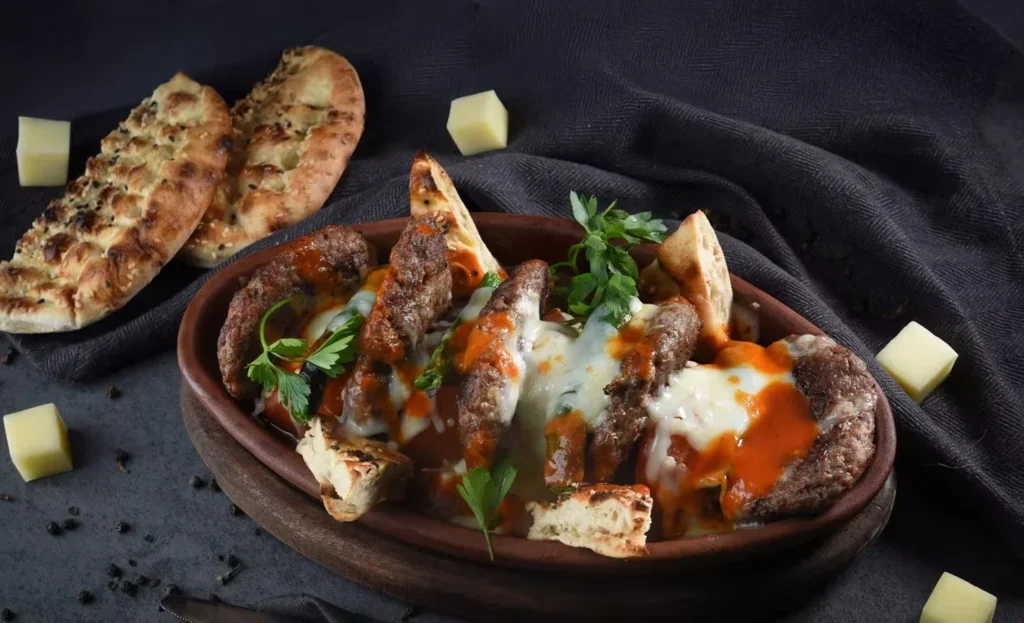 Late Night Food in Dubai: A dish featuring grilled meat patties topped with melted cheese and tomato sauce, garnished with parsley, and served with pieces of pita bread on a dark surface with cheese cubes nearby.