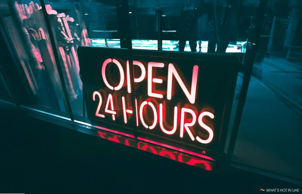 A neon sign reading "OPEN 24 HOURS" in bright red letters, illuminated against a dark background with reflections in a window.