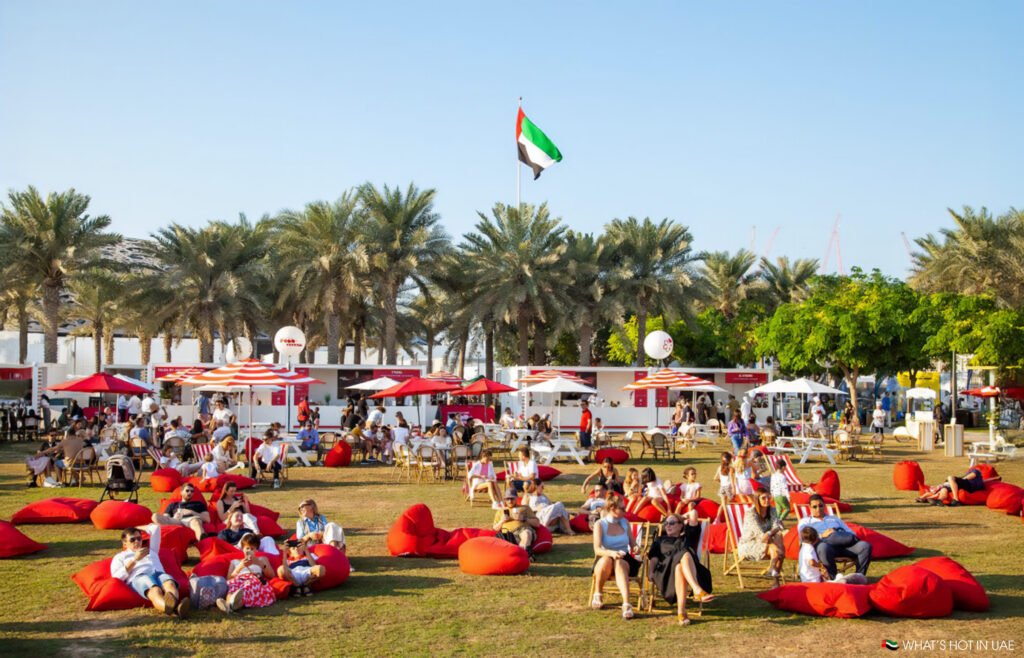 People relaxing on red bean bags and chairs in an outdoor area with palm trees, surrounded by food stalls with umbrellas, under a clear blue sky and flying a flag with the design of the United Arab Emirates.