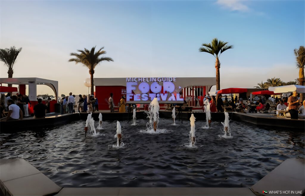 A bustling outdoor scene of the Michelin Guide Food Festival with a fountain in the foreground, people mingling around outdoor seating, and palm trees under a clear sky.