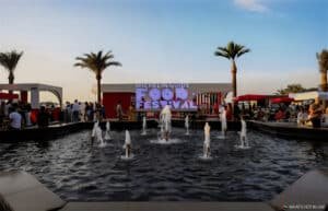 A bustling outdoor scene of the Michelin Guide Food Festival with a fountain in the foreground, people mingling around outdoor seating, and palm trees under a clear sky.