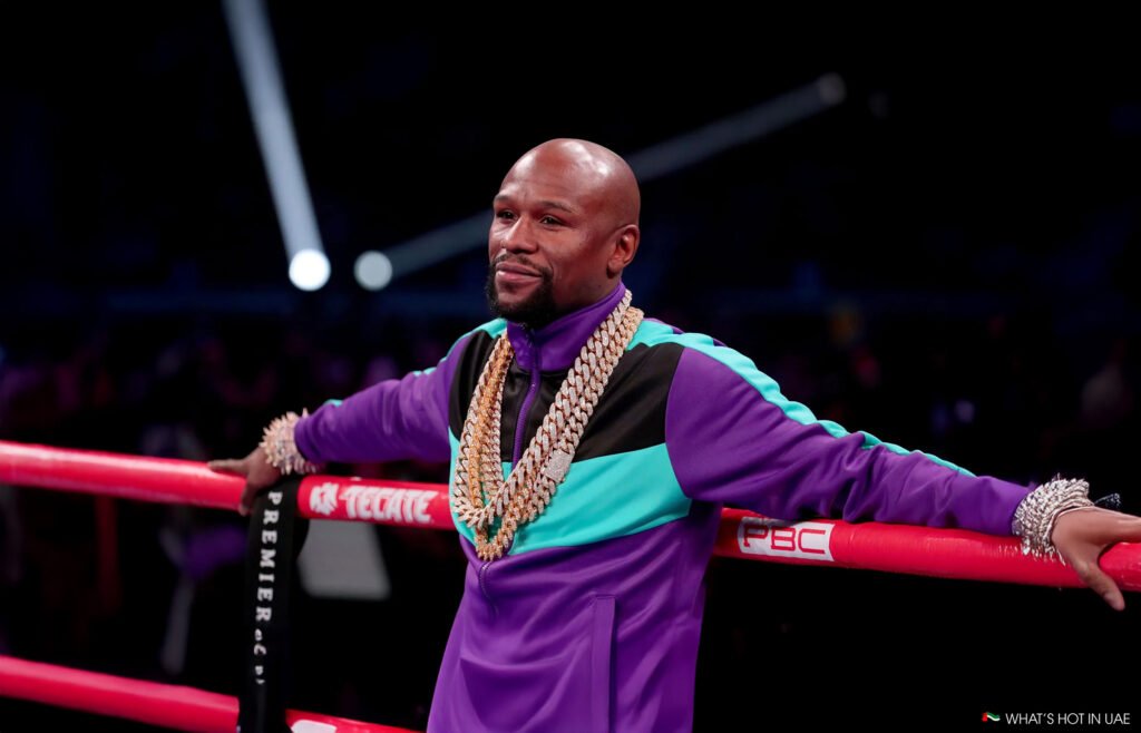 Pacquiao vs. Mayweather Rematch: A man in a purple and turquoise tracksuit stands in a boxing ring, adorned with large gold chains around his neck and diamond bracelets on his wrist.