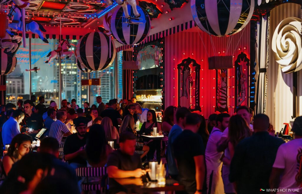 A vibrant indoor bar scene with people socializing under red and blue lighting, surrounded by whimsical decor such as striped balloons and carousel horses.