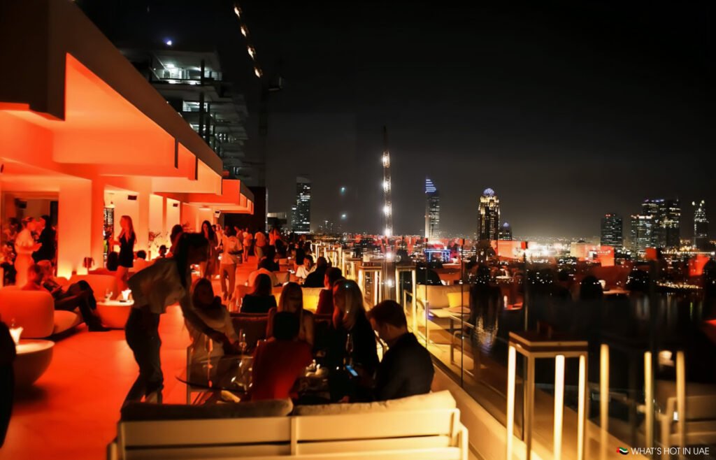 A rooftop bar bustling with people at night, illuminated by warm orange lighting, with a city skyline visible in the background.