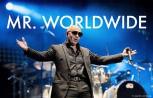 Pitbull in a dark suit and sunglasses is performing on stage with his arms outstretched, holding a microphone. The text "MR. WORLDWIDE" is displayed above him. The background features a blue-lit stage with musical instruments visible.