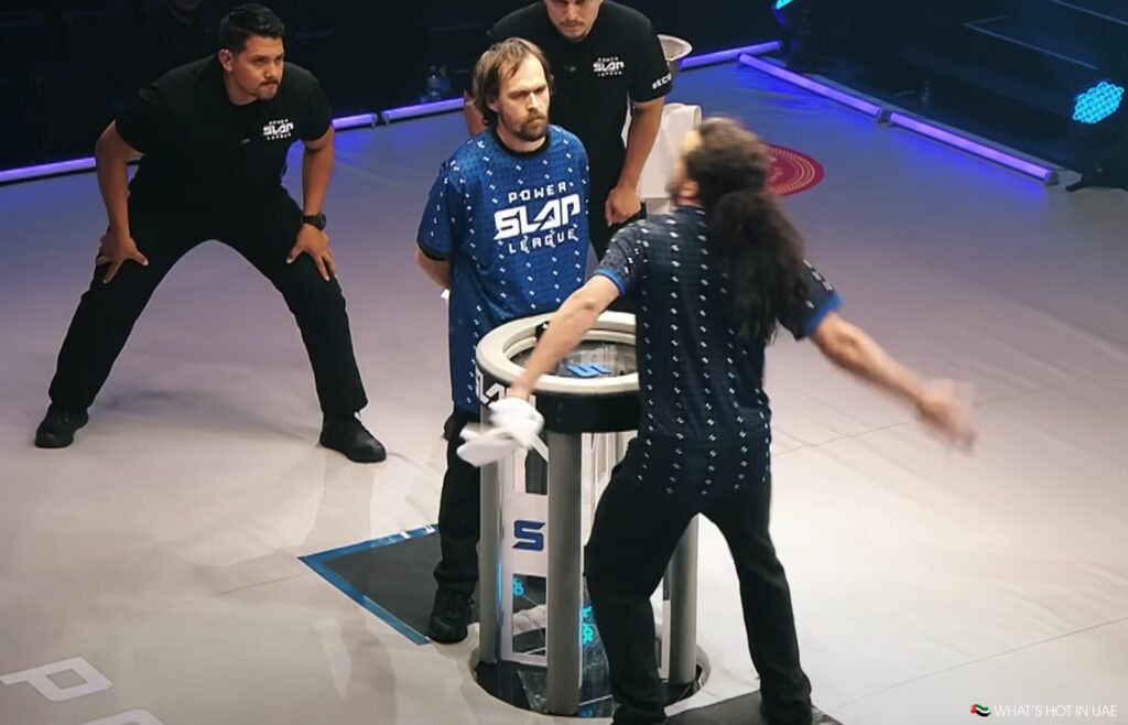 Two participants in blue jerseys face each other over a round table in a slap competition setting, with one person preparing to slap while the other stands with hands behind back. Two referees in black shirts observe closely.