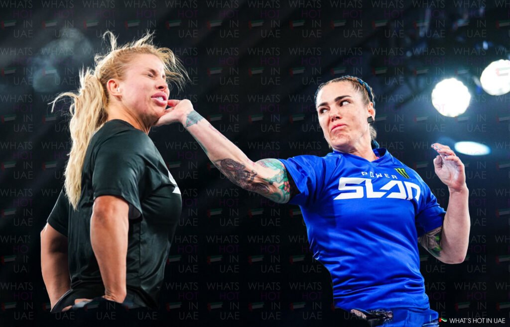 Two women in an intense slap match, one wearing a black shirt being slapped by the other in a blue shirt with tattoos on her arm, in front of a background with 'What's Hot in UAE' logo.