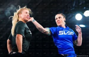Two women in an intense slap match, one wearing a black shirt being slapped by the other in a blue shirt with tattoos on her arm, in front of a background with 'What's Hot in UAE' logo.