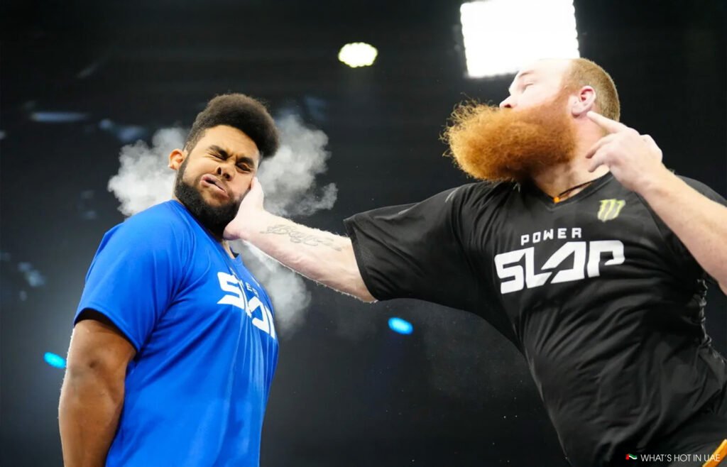 Power Slap UAE: A man in a blue shirt is being slapped by another man with a red beard wearing a black "Power Slap" shirt, with a cloud of impact visible from the slap.