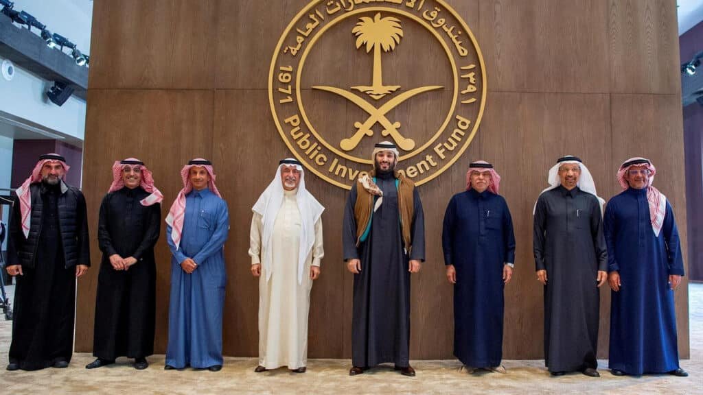 A group of men in traditional Middle Eastern attire stand in front of a wooden wall with the Public Investment Fund emblem, featuring a stylized palm tree and crossed swords, inscribed in both Arabic and English.