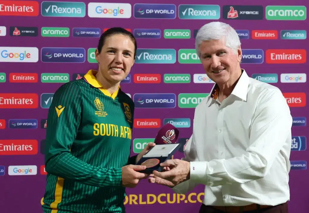 A person wearing a green South Africa cricket jersey receives an award from another person in a white shirt. The backdrop displays logos from sponsors including Emirates, Google, and Rexona, indicating a cricket event. South Africa World Cup win.