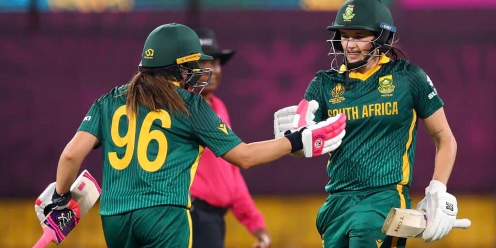 Two South African women cricketers wearing green uniforms with yellow accents, high-fiving each other on the field during a match, with one wearing a helmet and both holding cricket bats.