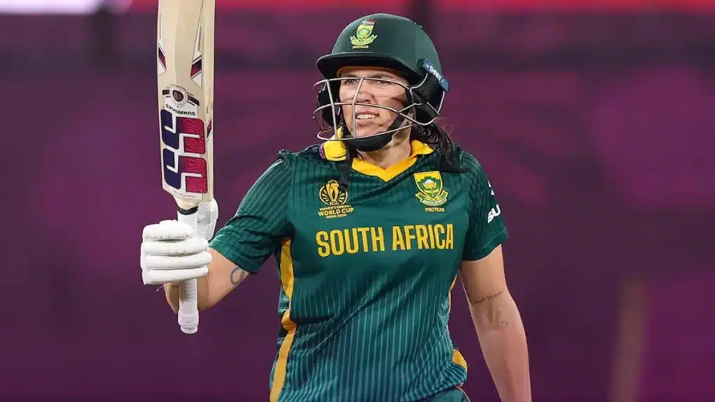 A South African female cricket player wearing a green and yellow uniform holds up her bat in celebration on the field. She is wearing a helmet with a visor and gloves, with the word 'PROTEAS' and the team logo visible on her jersey.