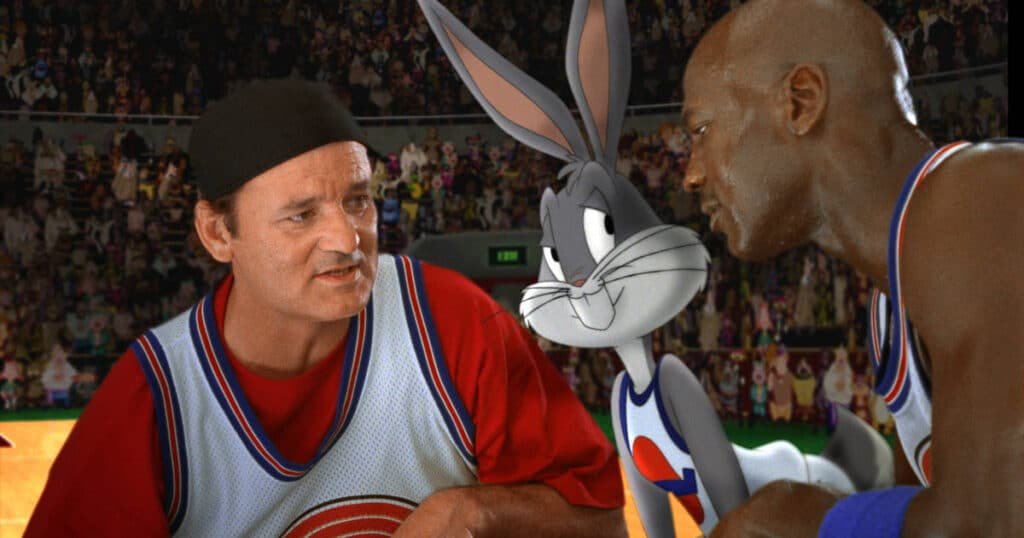 A basketball scene from the movie Space Jam featuring a cartoon rabbit character and two men in jerseys talking on a court, with a crowded stadium in the background.