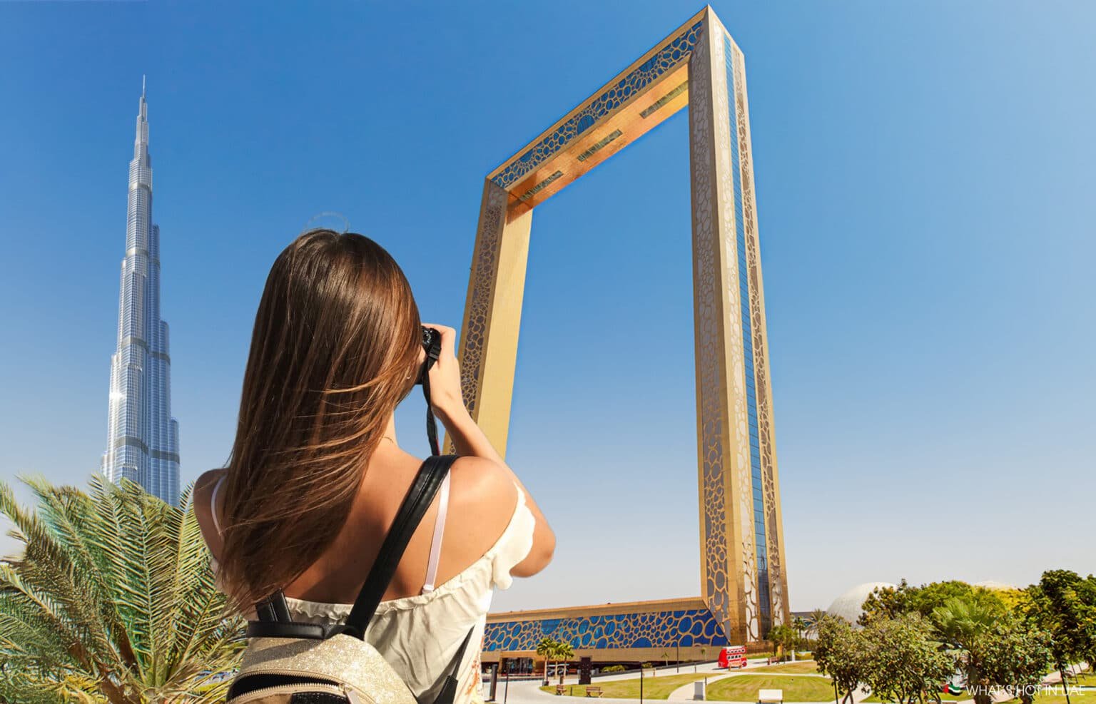 A woman with long brown hair is taking a photo of the Dubai Frame with a camera. The Burj Khalifa is visible in the background against a clear blue sky.