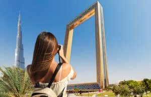 A woman with long brown hair is taking a photo of the Dubai Frame with a camera. The Burj Khalifa is visible in the background against a clear blue sky.
