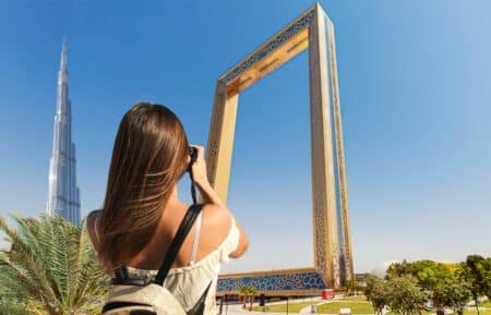 A woman with long brown hair is taking a photo of the Dubai Frame with a camera. The Burj Khalifa is visible in the background against a clear blue sky.