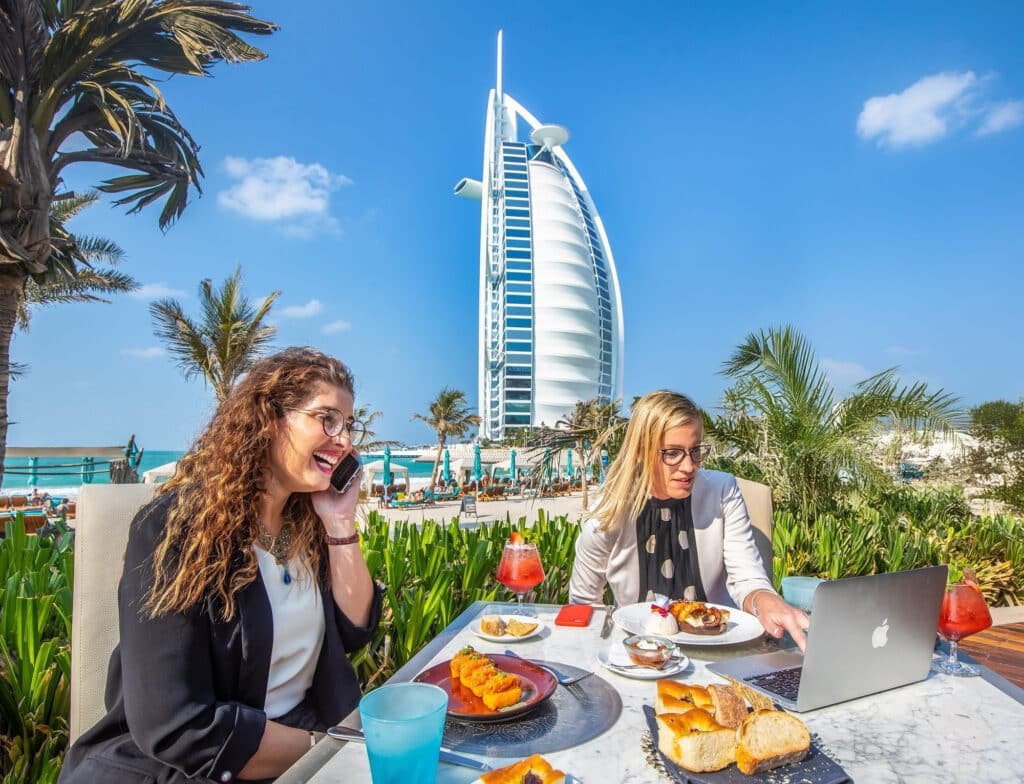 Two women sitting at an outdoor cafe table with plates of food, one talking on the phone, and the other working on a laptop, with the iconic sail-shaped Burj Al Arab hotel in the background, surrounded by palm trees and a clear blue sky.